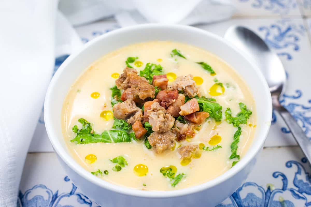 A bowl of Creamy Tuscan Soup with Sausage and Kale set on a blue and white tiled surface with a spoon and napkin beside it.