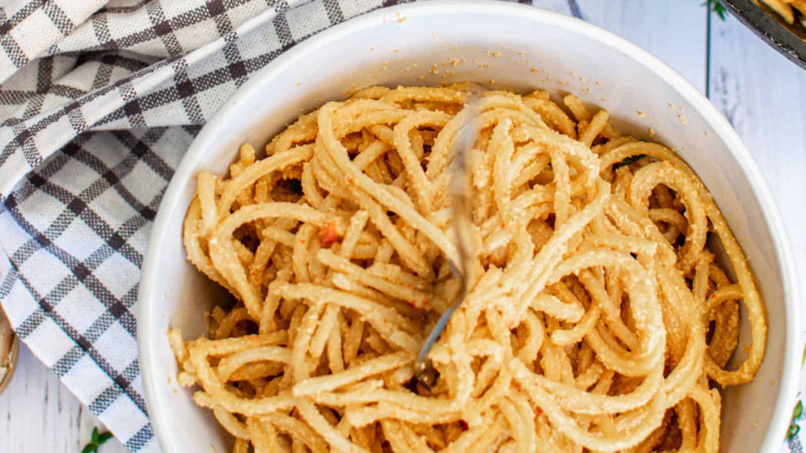 A bowl of Creamy Vegan Red Pepper Pasta with a fork twirled into the noodles, served on a white wooden surface with a checkered napkin beside it.