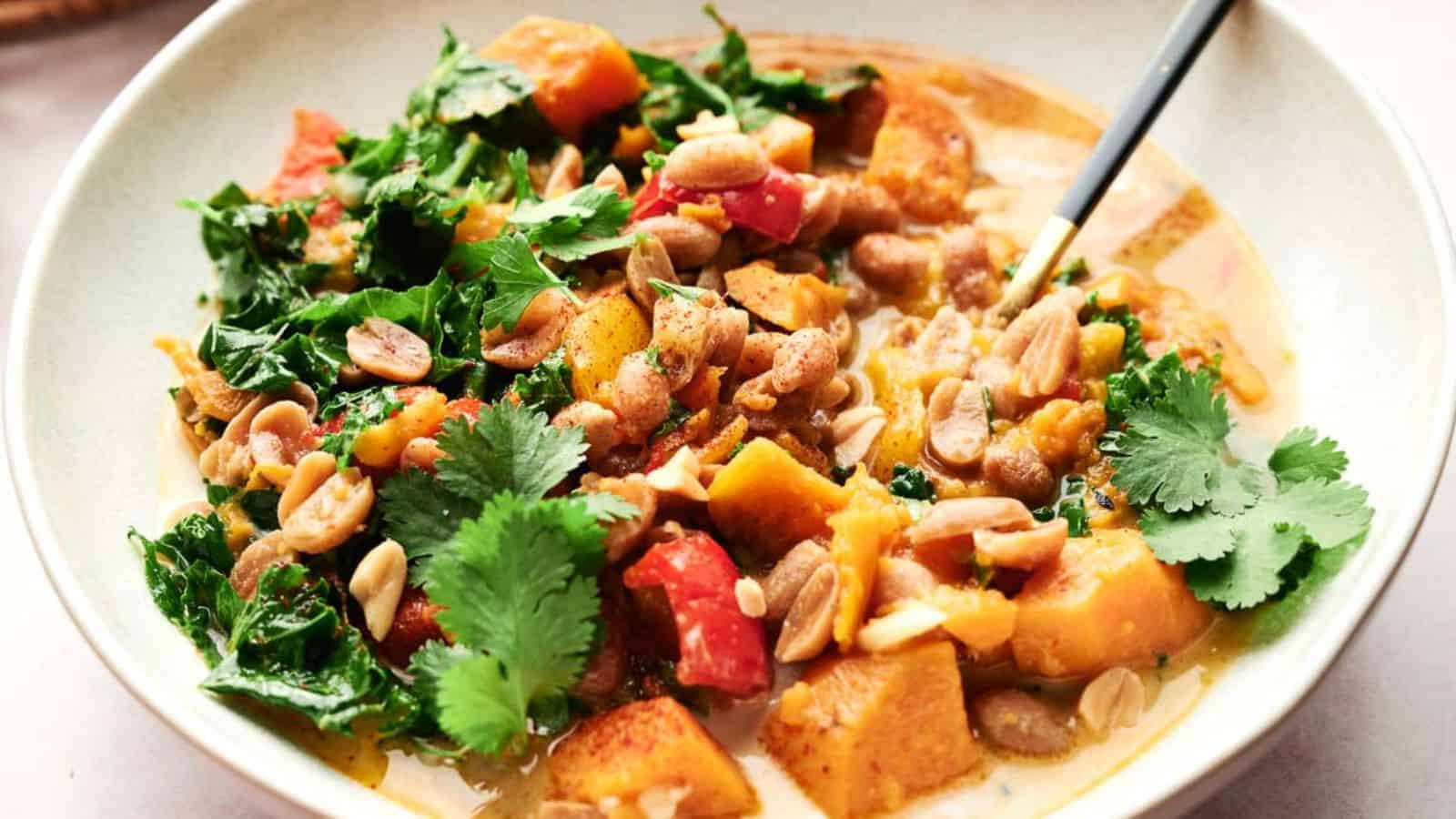 A bowl of vegetable curry with leafy greens, chunks of squash, diced tomatoes, peanuts, and cilantro, served with a spoon.