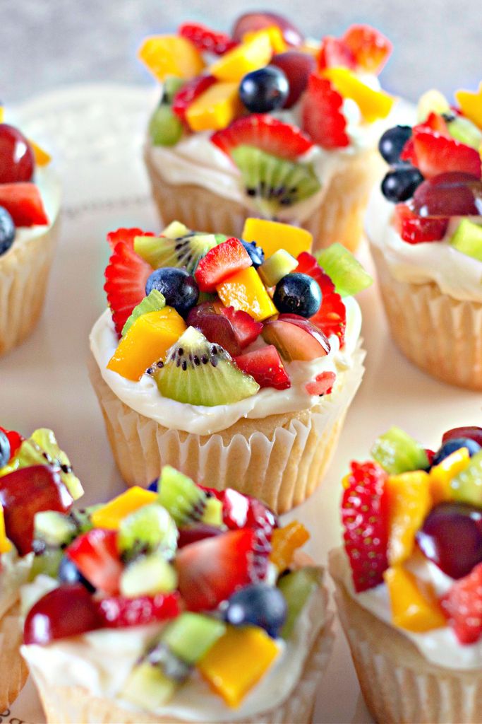 “Fruit Pizza” Cupcakes with a creamy white frosting, topped with fresh fruit including strawberries, blueberries, kiwi, mango, and grapes, arranged on a white plate.
