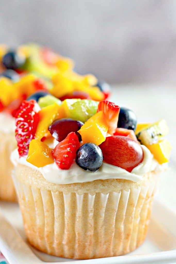A “Fruit Pizza” Cupcakes with creamy white frosting, topped with fresh fruit including strawberries, blueberries, kiwi, mango, and grapes, sitting on a white plate.