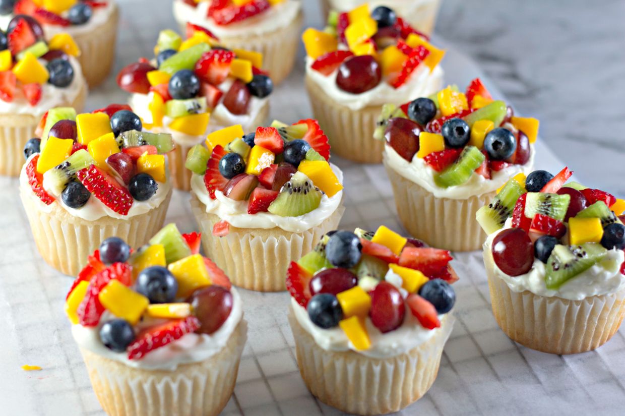 “Fruit Pizza” Cupcakes topped with a layer of creamy white frosting and an assortment of fresh fruit, including strawberries, blueberries, kiwi, mango, and grapes, arranged on a marble surface.