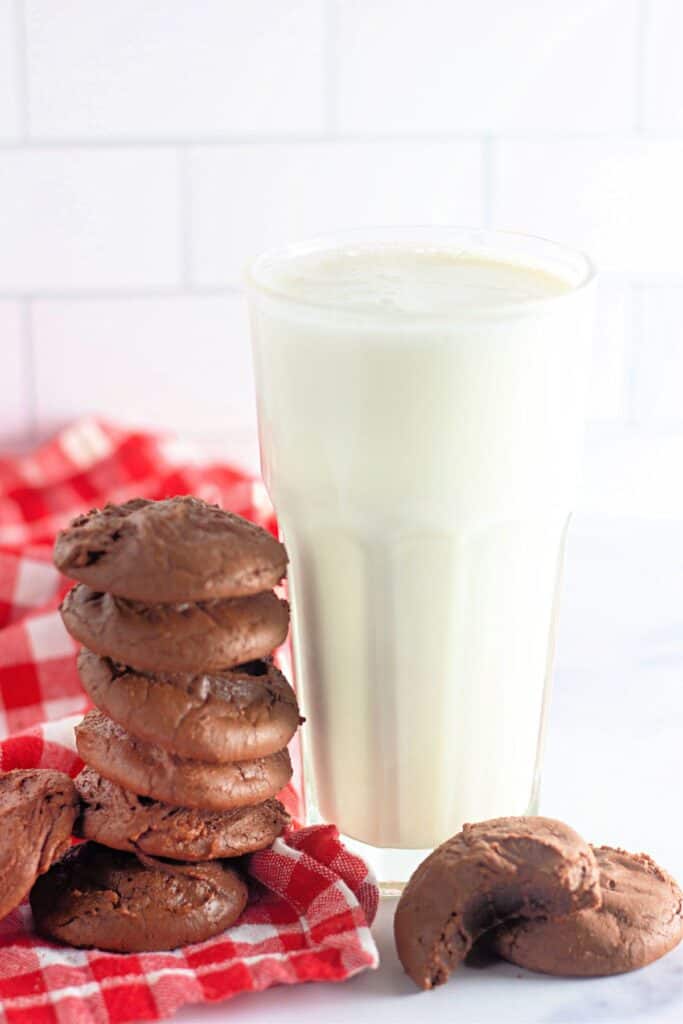 A stack of fudgy chocolate brownie cookies next to a glass of milk, placed on a red and white checkered cloth.