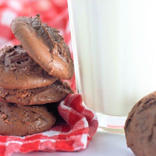 Stack of fudgy chocolate brownie cookies next to a glass of milk, placed on a red and white checkered cloth.