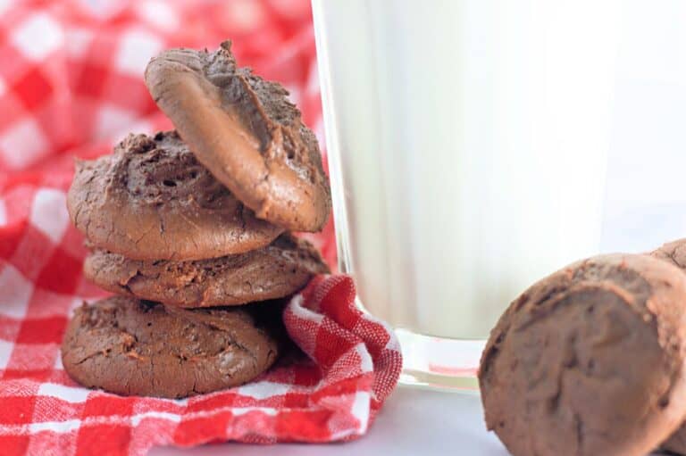 Stack of fudgy chocolate brownie cookies next to a glass of milk, placed on a red and white checkered cloth.