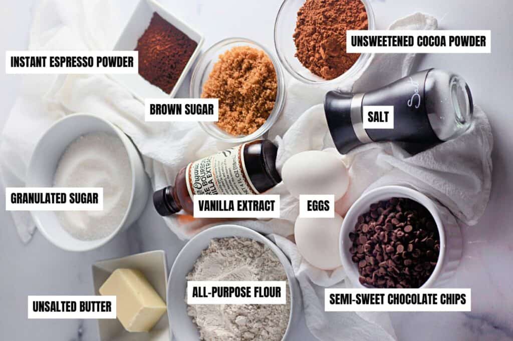 Various ingredients for fudgy chocolate brownie cookies, including instant espresso powder, brown sugar, granulated sugar, vanilla extract, eggs, unsweetened cocoa powder, salt, all-purpose flour, unsalted butter, and semi-sweet chocolate chips.