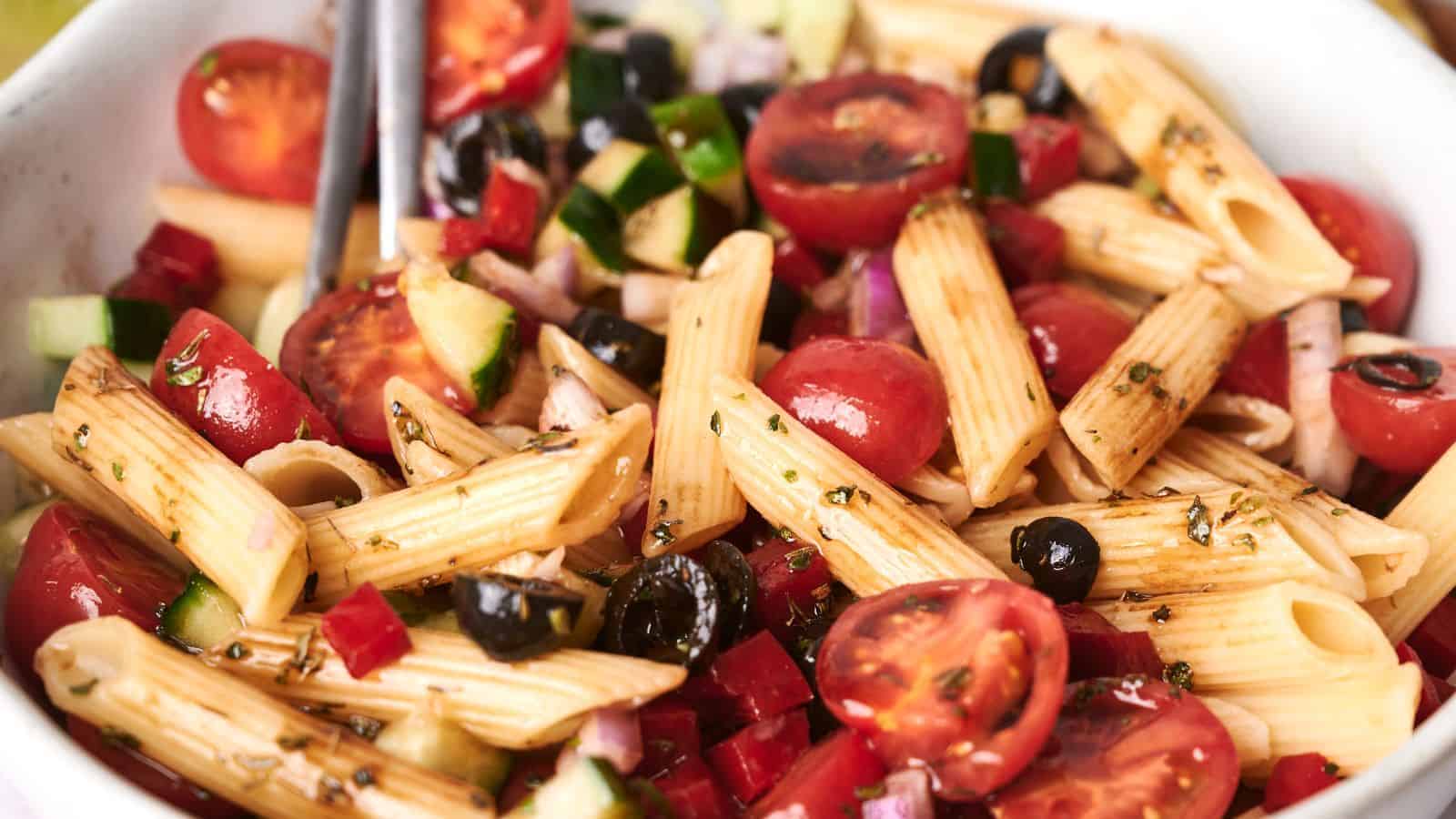 A close-up of a pasta salad with penne, cherry tomatoes, cucumbers, black olives, red onions, and seasonings in a white bowl.