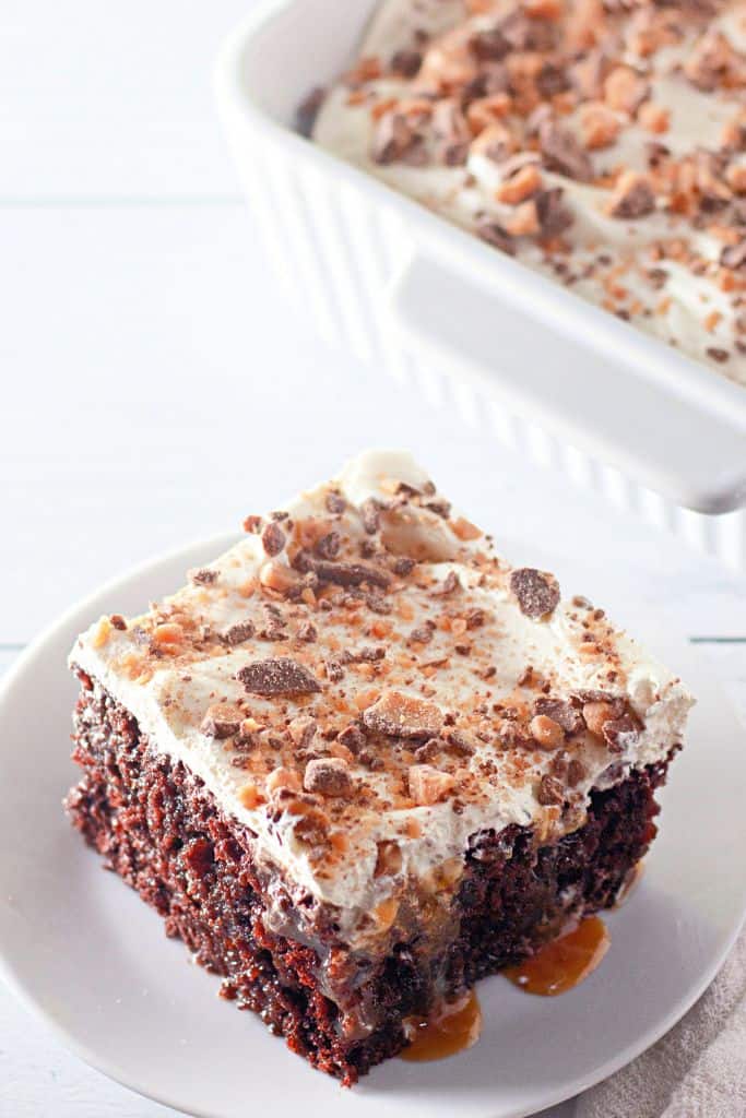 A close-up of a slice of Heath Bar poke cake on a white plate, showing moist chocolate cake, caramel drizzle, whipped topping, and crushed toffee bits.