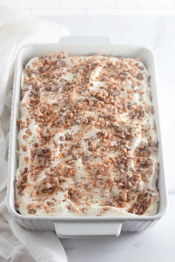 A full view of a Heath Bar poke cake in a white baking dish, topped with whipped cream and crushed toffee bits.