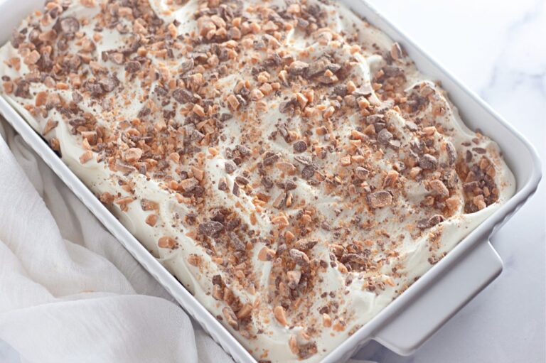 A close-up of a Heath Bar poke cake in a white baking dish, topped with whipped cream and crushed toffee bits.