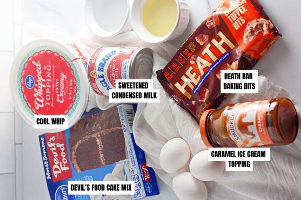 An overhead view of ingredients for a Heath Bar poke cake, including cake mix, Cool Whip, sweetened condensed milk, caramel topping, Heath Bar baking bits, eggs, and oil.