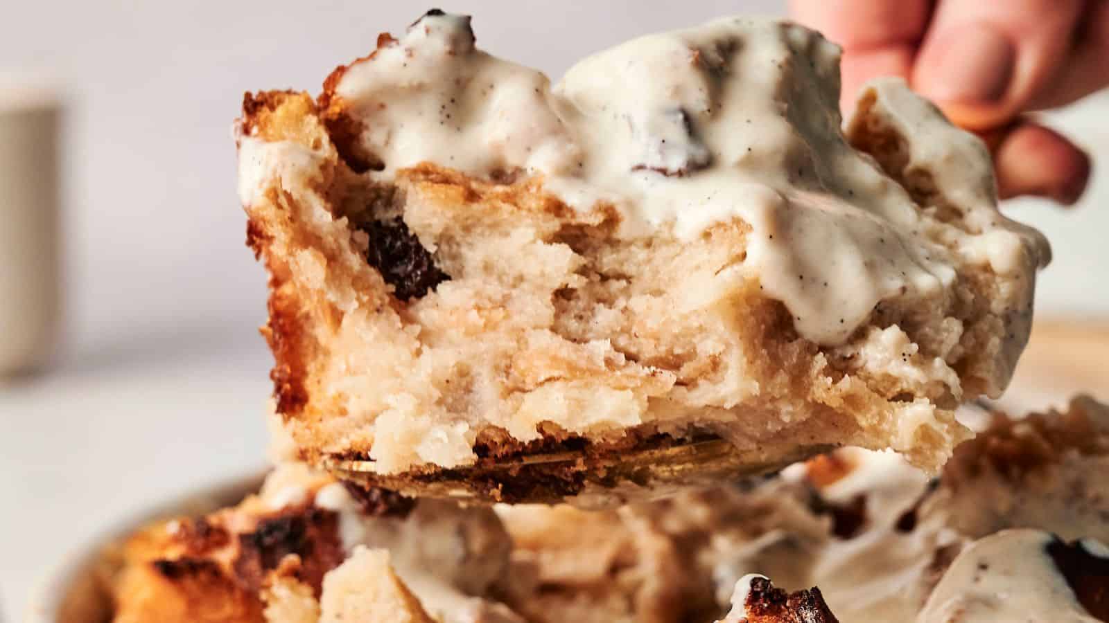 A close-up of decadent bread pudding, topped with creamy vanilla sauce, is being lifted from a serving plate.