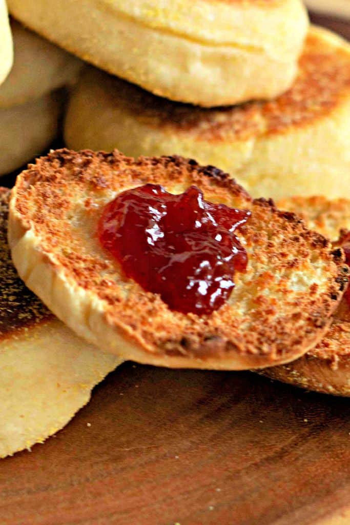A toasted homemade English muffin topped with glossy red jam, sitting on a wooden surface with more muffins in the background.