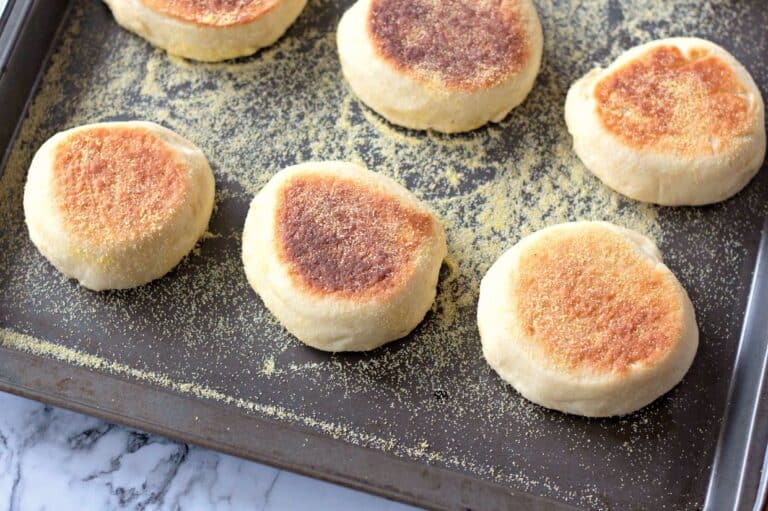 Freshly baked homemade English muffins on a baking sheet, dusted with cornmeal. The muffins have a golden-brown crust on top and a soft, fluffy texture.