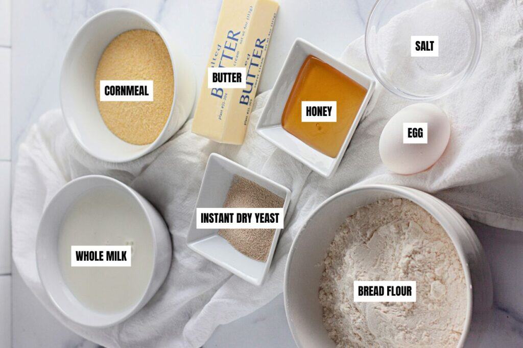Labeled ingredients for homemade English muffins, including cornmeal, butter, honey, salt, egg, instant dry yeast, whole milk, and bread flour.