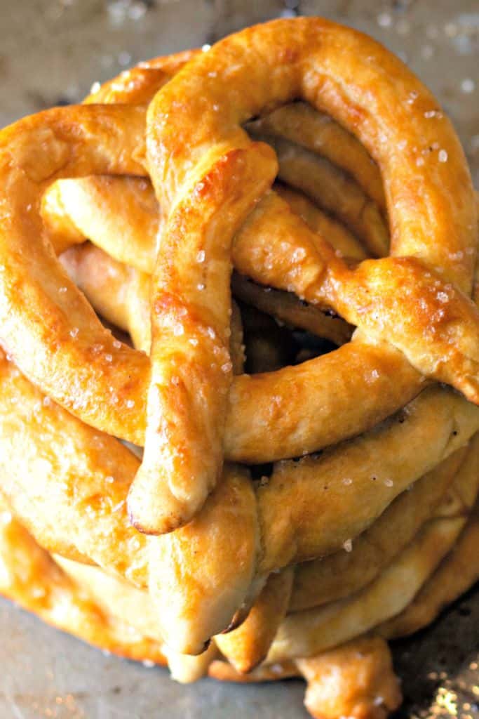 A stack of golden-brown homemade soft pretzels with a glossy surface and coarse salt sprinkled on top, resting on a baking sheet.