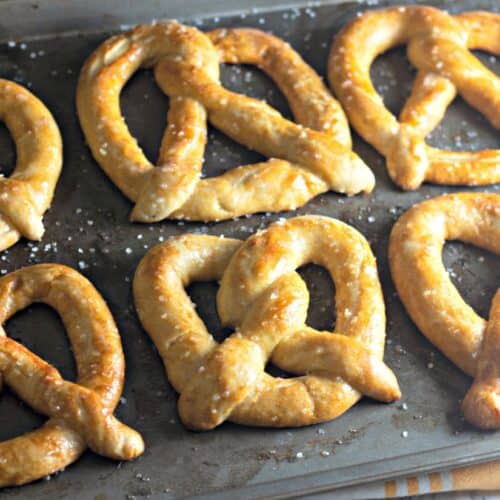 A baking sheet filled with six golden-brown homemade soft pretzels, sprinkled with coarse salt.
