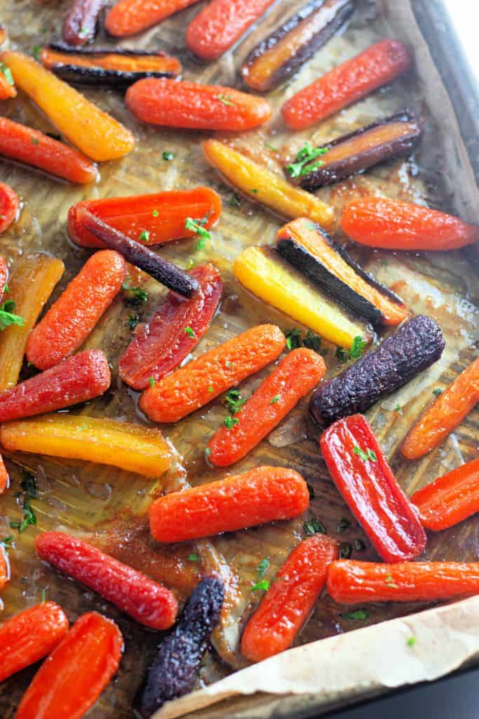 A baking sheet lined with parchment paper holds colorful baby carrots roasted to a caramelized finish, glistening with honey glaze and garnished with finely chopped herbs.