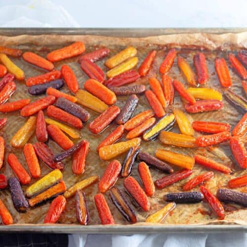 A baking sheet lined with parchment paper holds honey-roasted baby carrots in vibrant orange, yellow, and purple hues. The carrots are glistening with a caramelized glaze.