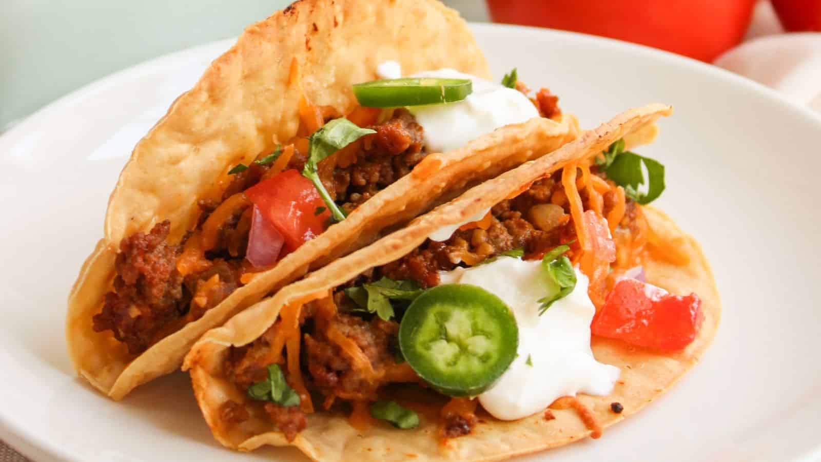 Two tacos with beef, diced tomatoes, jalapeño slices, shredded cheese, sour cream, and cilantro on a white plate.