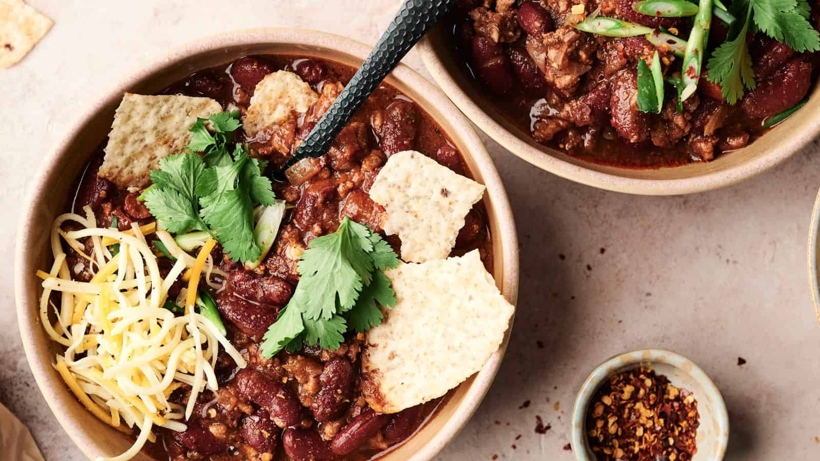 Two bowls of chili with kidney beans, meat, tortilla chips, shredded cheese, and cilantro. A black spoon in one bowl. Additional chili flakes in a small dish nearby.