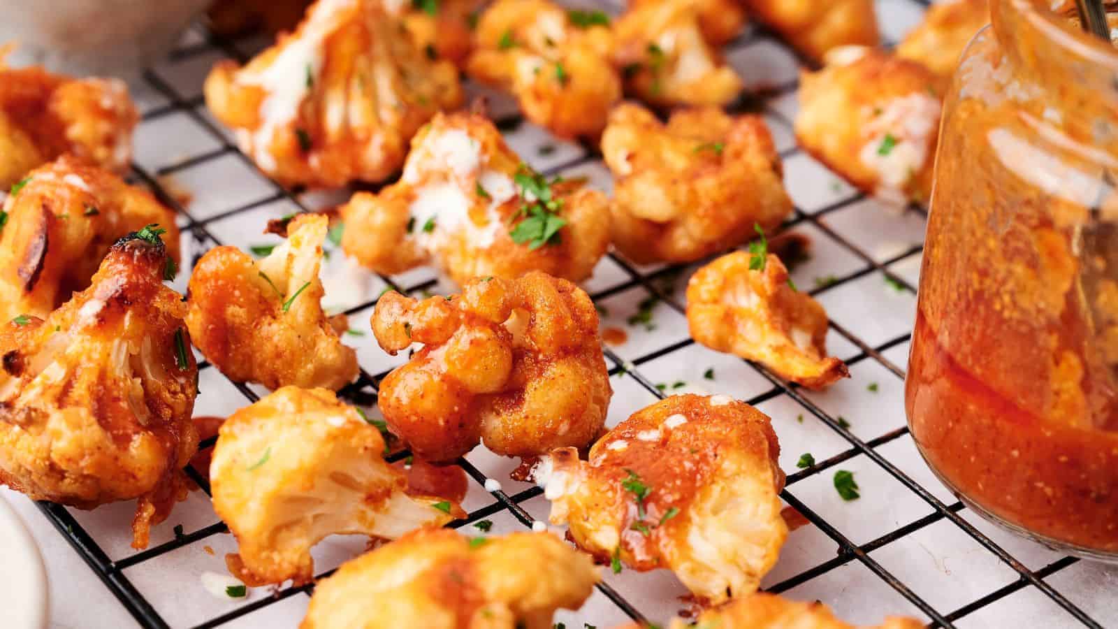 Close-up of baked Buffalo Cauliflower Wings coated in orange sauce, scattered on a cooling rack, with a glass jar of sauce beside them. Garnished with chopped herbs.