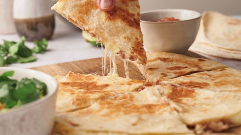 A hand lifts a cheesy quesadilla slice from a wooden board. Bowls with salsa and cilantro are in the background.