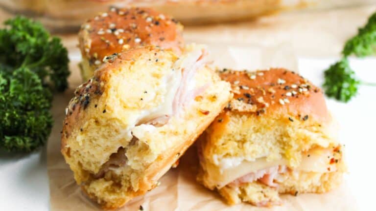 Three ham and cheese sliders on seeded buns are on display, surrounded by fresh parsley.
