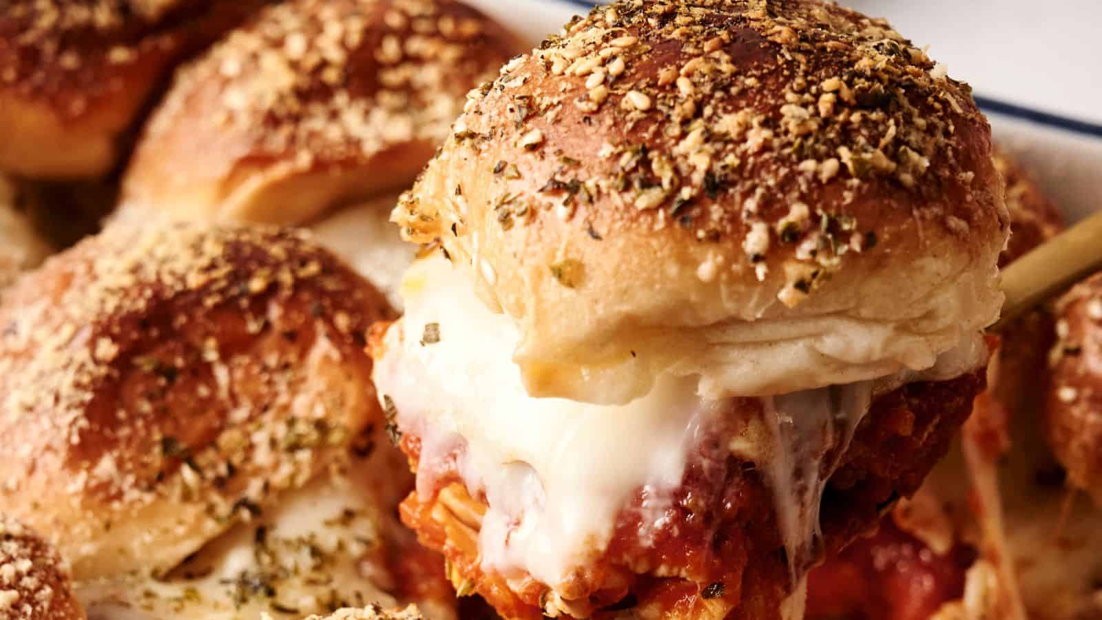 Close-up of a cheesy meatball slider with a seasoned bun, showing melted cheese and tomato sauce. Other sliders are visible in the background.