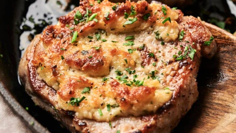 Close-up of a cooked pork chop topped with melted cheese and garnished with chopped parsley, served on a dark plate.