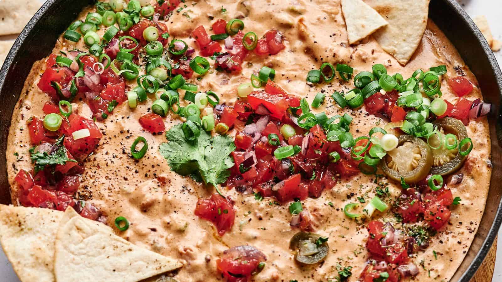 A skillet filled with creamy cheese dip topped with chopped tomatoes, green onions, cilantro, and sliced jalapeños, surrounded by triangular tortilla chips.