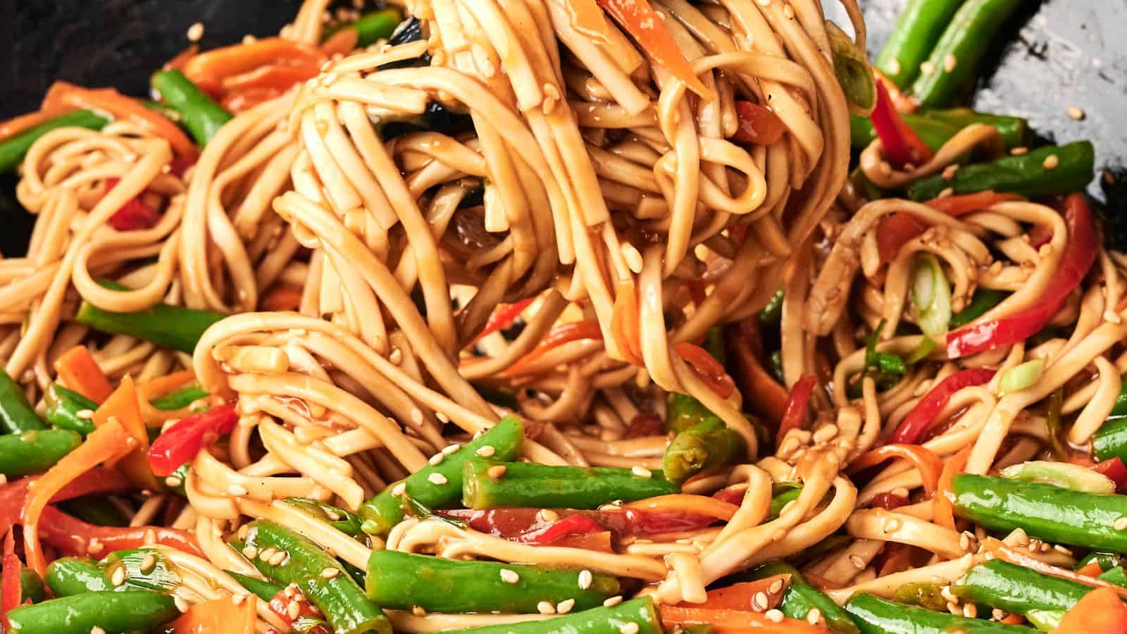 A close-up of stir-fried noodles with green beans, sliced carrots, and bell peppers, topped with sesame seeds.
