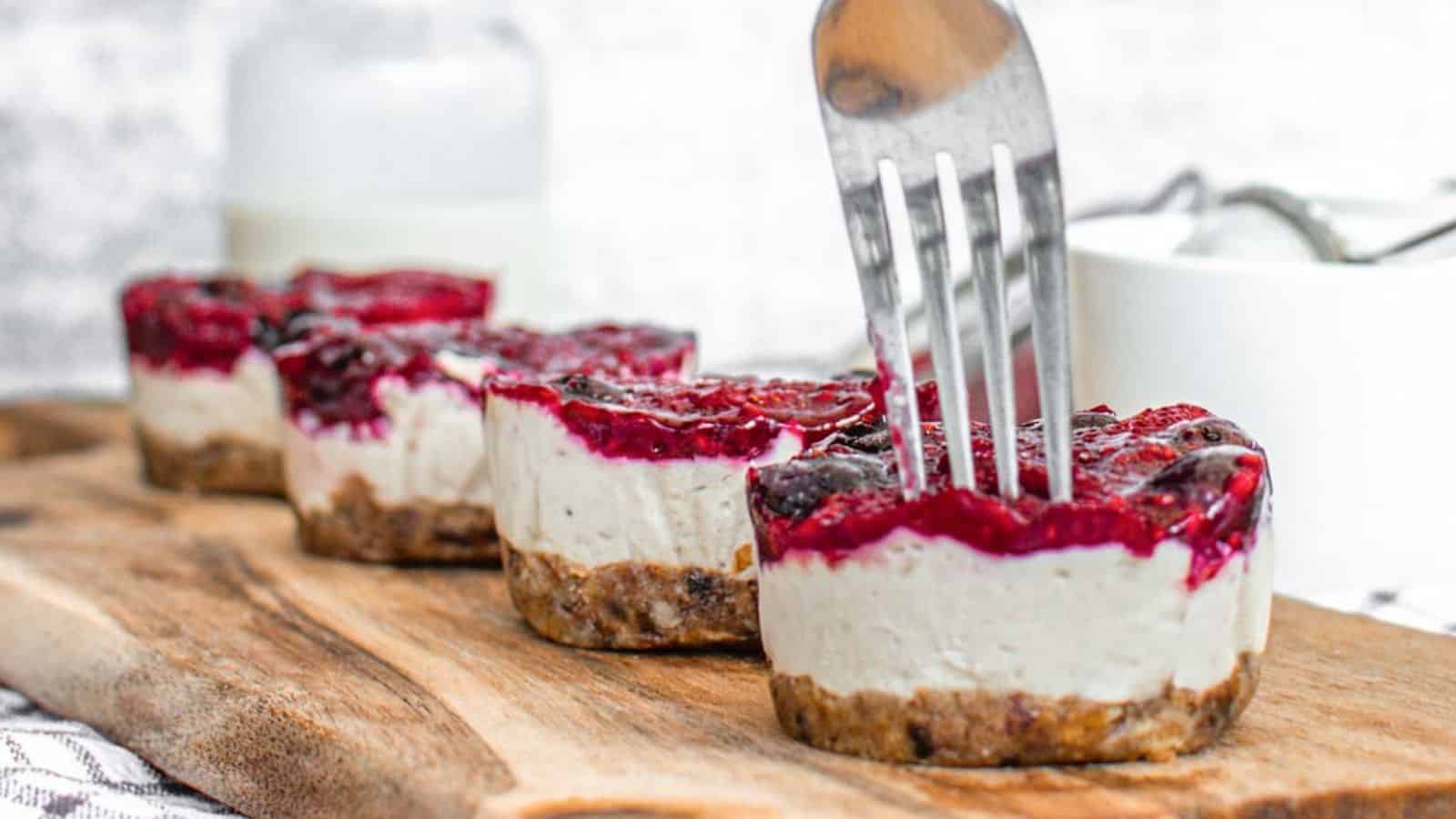 No-bake mini vegan cheesecakes with a berry topping on a wooden board, with a fork cutting into one.