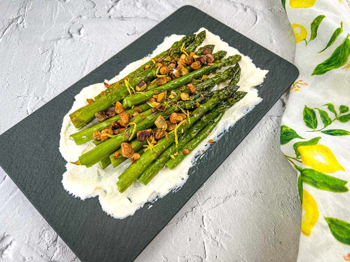 Oven-roasted asparagus on a bed of mascarpone, topped with toasted garlic, nuts, and lemon zest, served on a black slate platter.