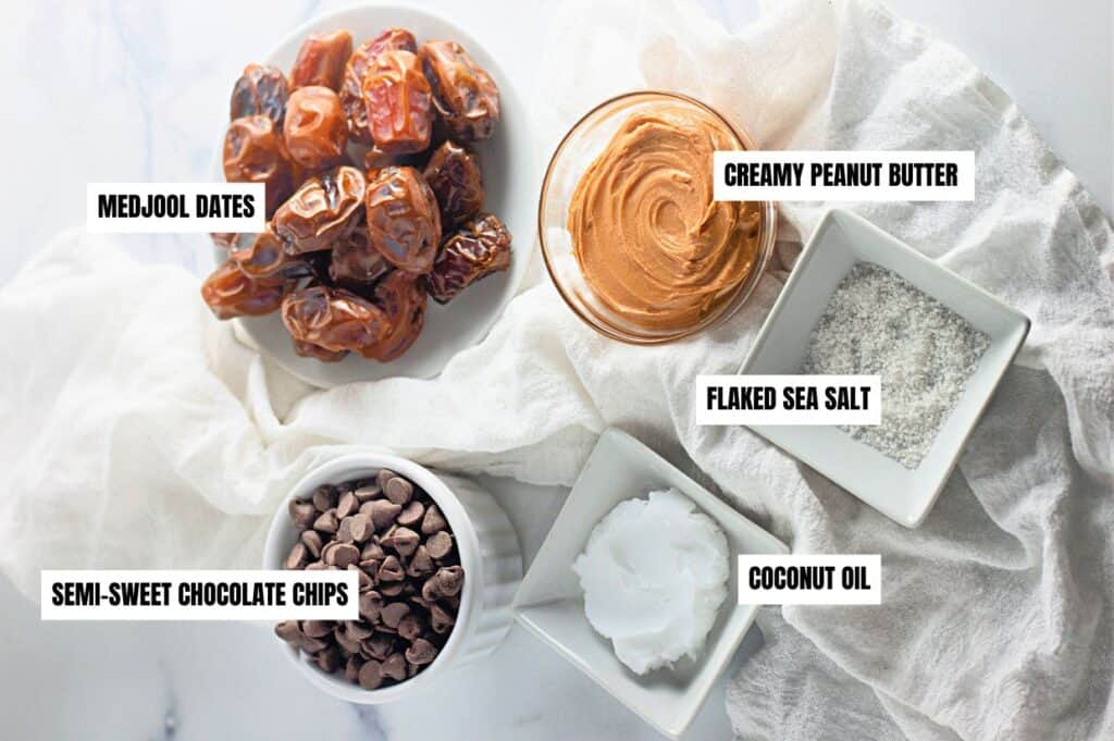 The image shows ingredients for a treat: Medjool dates, creamy peanut butter, flaked sea salt, semi-sweet chocolate chips, and coconut oil, arranged on a white cloth.