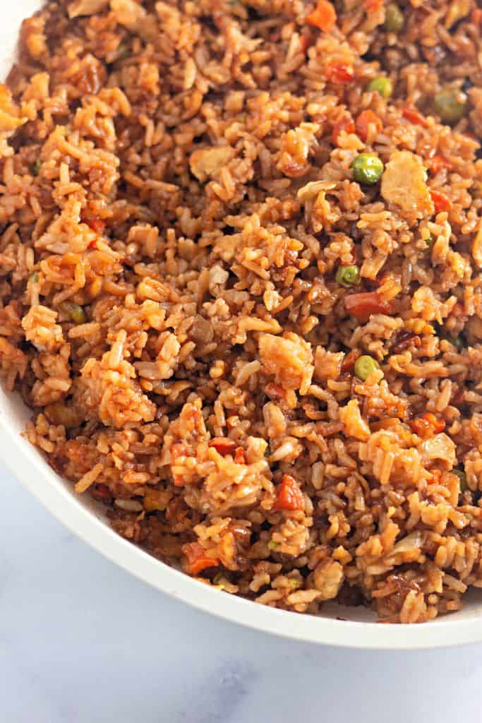 A close-up of a skillet filled with fried rice, featuring diced carrots, peas, scrambled egg, and chunks of chicken. The rice is coated in a dark sauce, giving it a rich, savory appearance.
