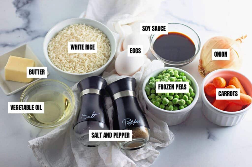 A top-down view of ingredients for making fried rice, each labeled with text. The ingredients include white rice, soy sauce, eggs, onion, frozen peas, carrots, butter, vegetable oil, and salt and pepper, arranged in bowls and containers on a marble surface.
