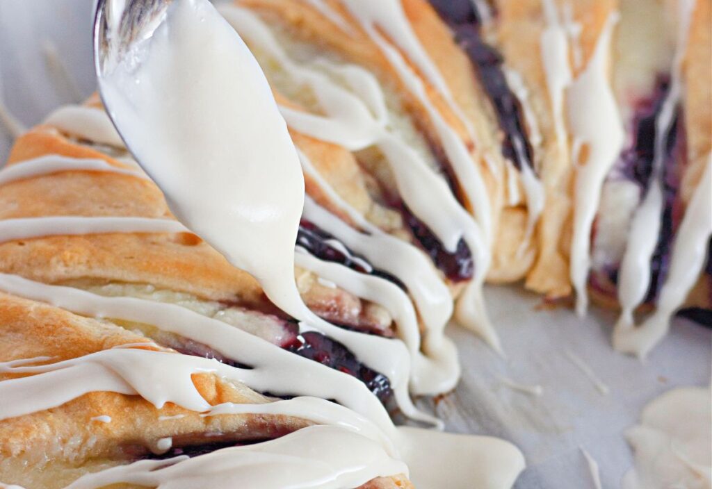 Close up of baked raspberry crescent ring drizzled with vanilla glaze, showing flaky layers and berry filling inside.