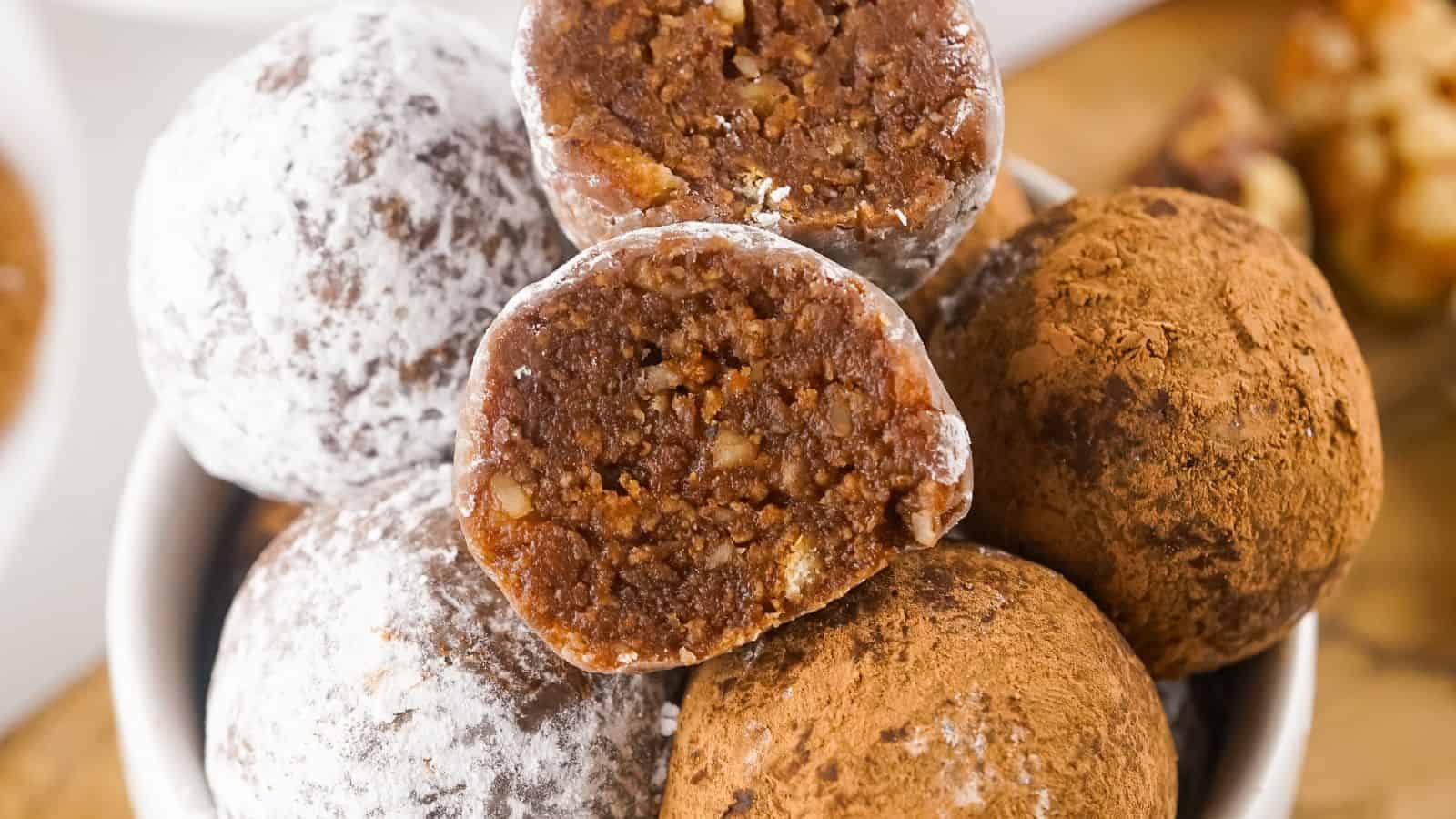 A bowl of assorted chocolate truffles coated in powdered sugar and cocoa powder, with one truffle split open to show the nutty interior.