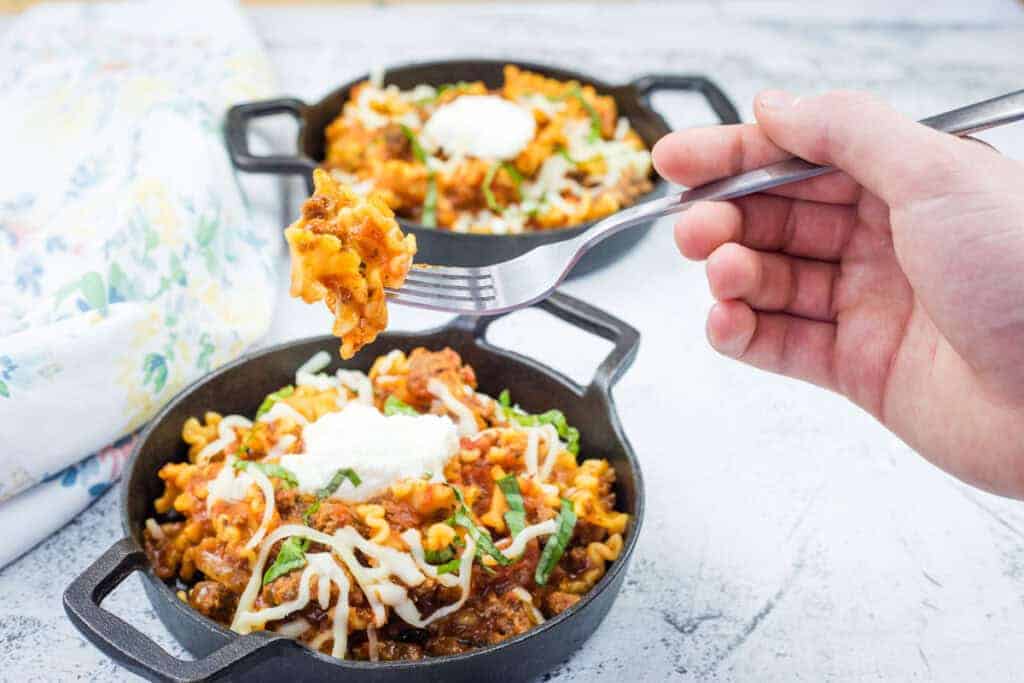 A hand holds a fork with a Classic Skillet Lasagna with Ground Beef topped with melted cheese and herbs.