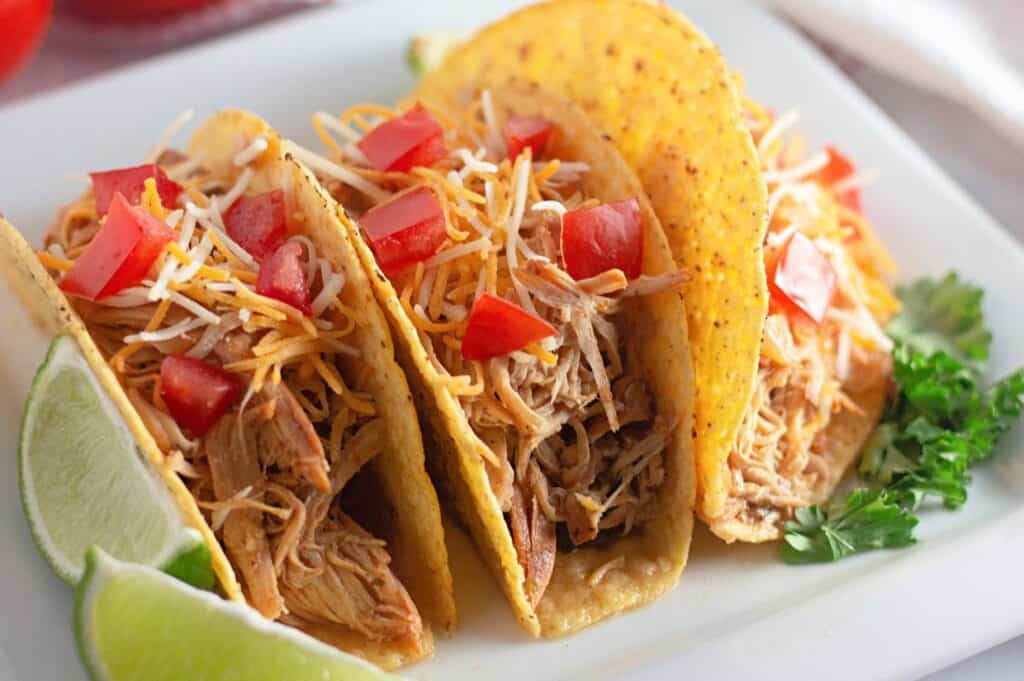 Close-up of three soft tacos filled with shredded chicken, topped with shredded cheese, diced tomatoes, and served with lime wedges and cilantro on the side.