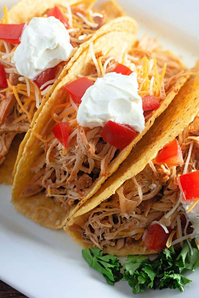 A three tacos filled with shredded chicken, topped with shredded cheese, diced tomatoes, and a dollop of sour cream.