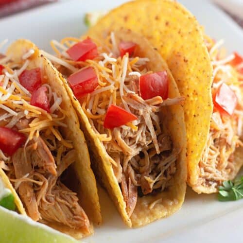 Close-up of three soft tacos filled with shredded chicken, topped with shredded cheese, diced tomatoes, and served with lime wedges and cilantro on the side.