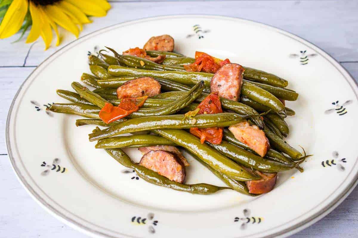A plate of cooked Southern Green Beans with sliced sausages and tomato pieces on a white dish, placed near a sunflower.