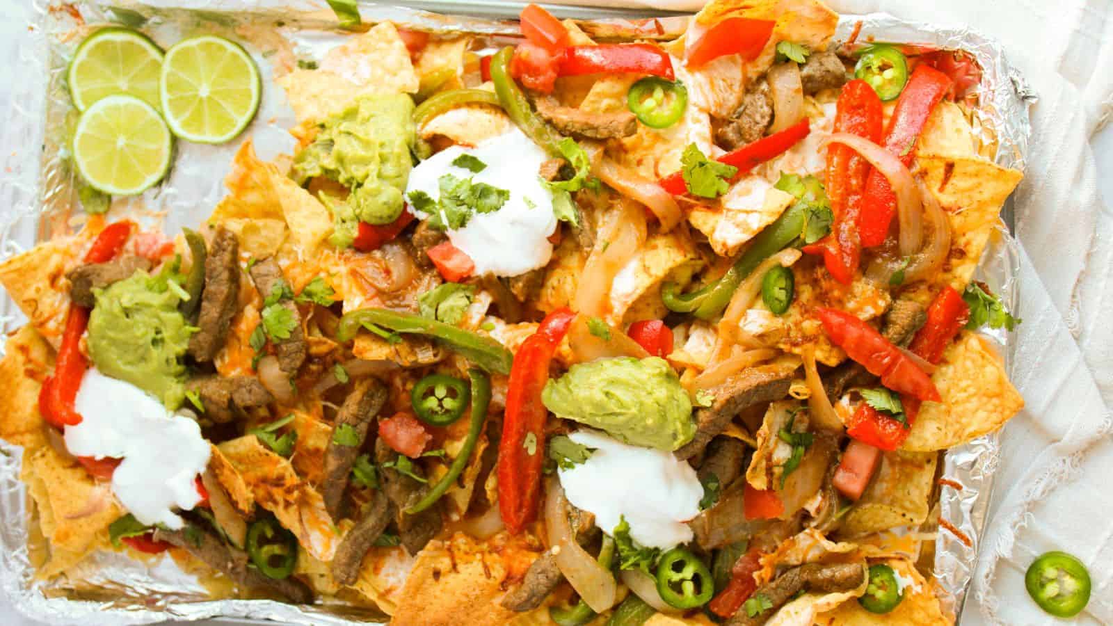 A tray of loaded nachos topped with sliced beef, red and green peppers, jalapeños, guacamole, sour cream, and lime wedges on the side.