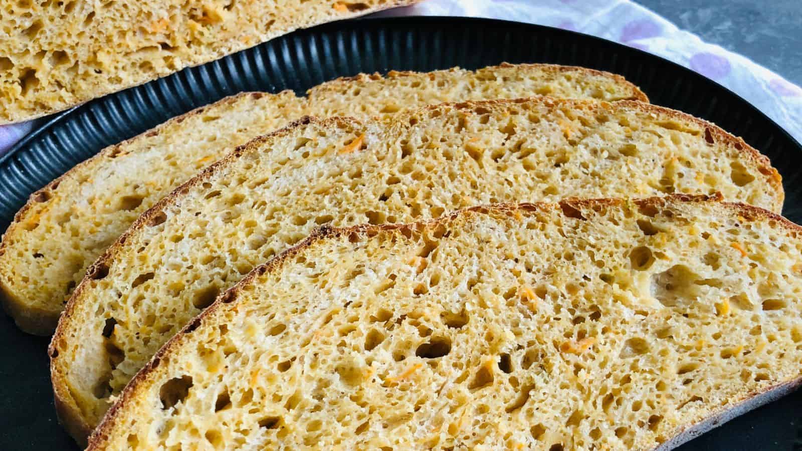 A black plate holds four slices of rustic bread with a golden crust and airy interior, with additional slices visible in the background.