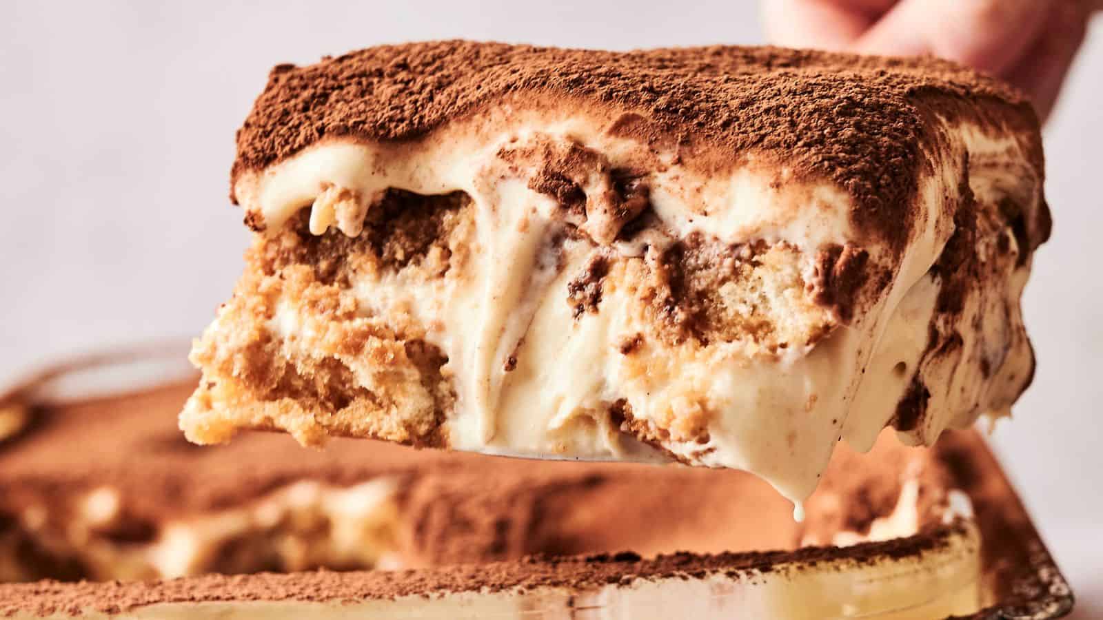 A slice of tiramisu is being lifted from the dish, showcasing layers of coffee-soaked ladyfingers and creamy mascarpone, topped with cocoa powder.