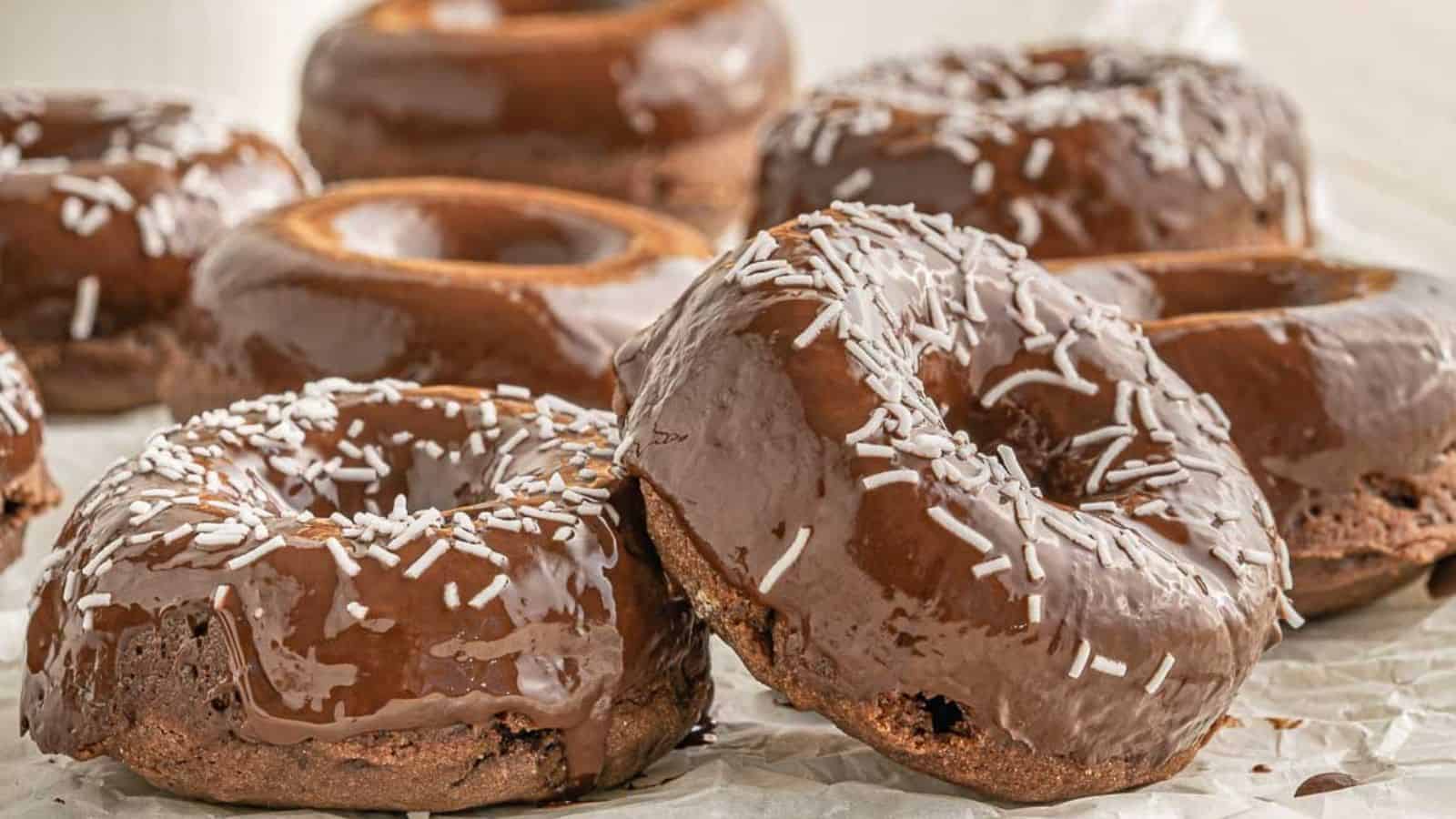 Close-up of vegan chocolate doughnuts with glossy chocolate glaze and white sprinkles, stacked on parchment paper.