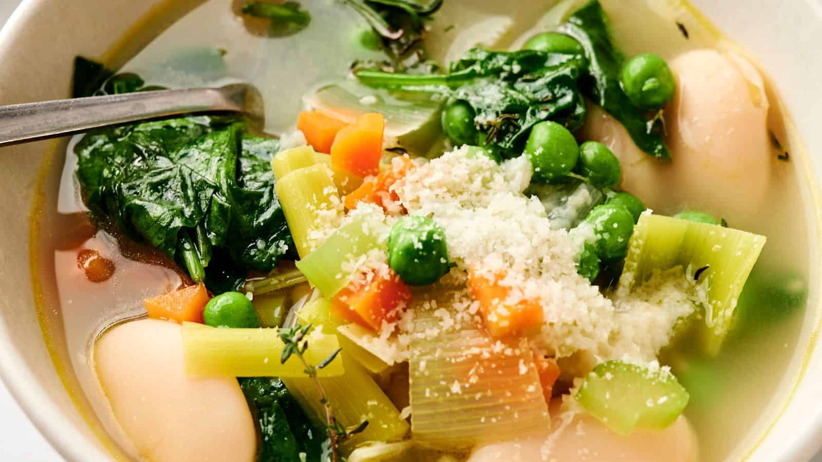 Close-up of a bowl of vegetable soup with spinach, peas, carrots, celery, and large beans, topped with grated cheese. A spoon rests in the bowl.