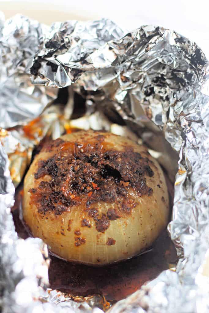 A baked onion wrapped in foil, with a flavorful, seasoned coating on top, and surrounded by a rich sauce. The onion is cooked until tender and caramelized.